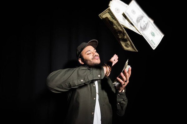 rich indian hip hop performer in cap throwing dollar banknotes on black 