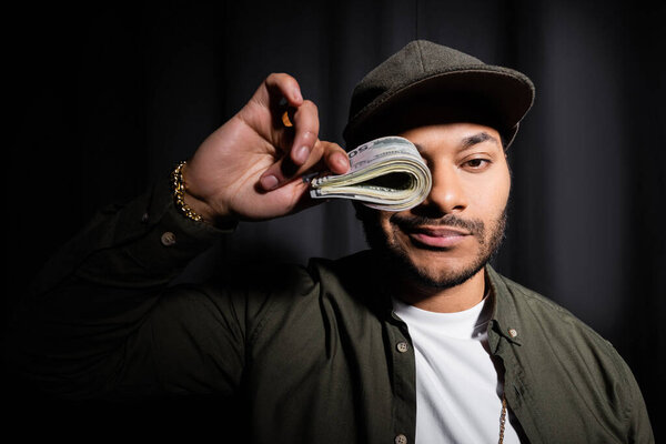 rich indian hip hop performer in cap covering eye with bundle of dollar banknotes on black 