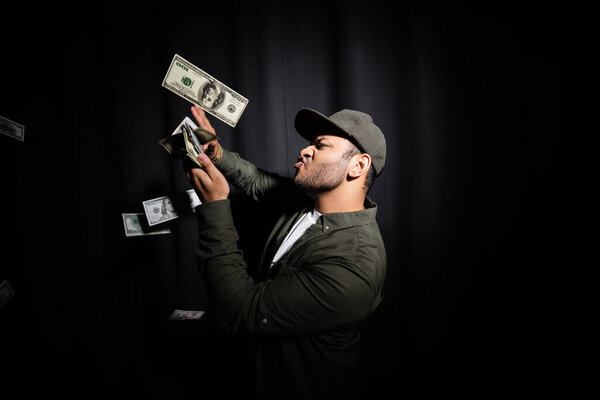 side view of rich indian hip hop performer in cap throwing dollar banknotes on black 