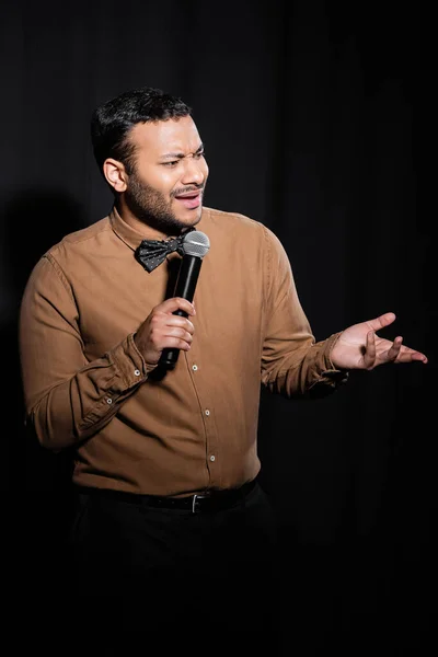 displeased indian stand up comedian in shirt and bow tie holding microphone during monologue on black