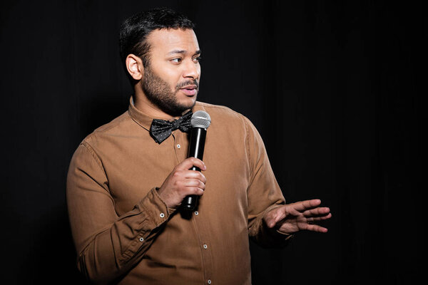 bearded indian stand up comedian in shirt and bow tie holding microphone during monologue on black 