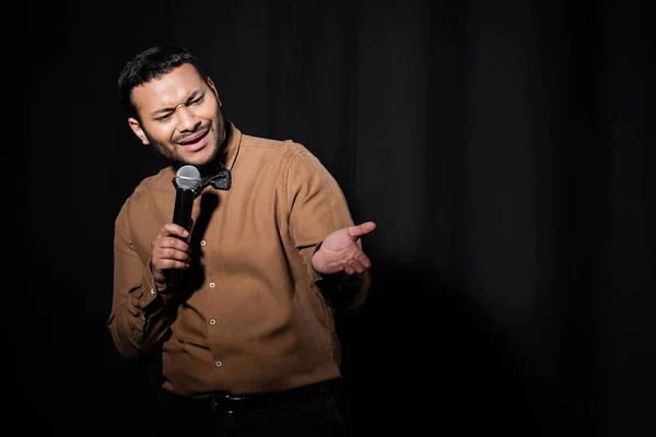 indian comedian in shirt and bow tie holding microphone and talking during performance on black