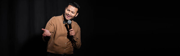 indian comedian in shirt and bow tie holding microphone and talking during performance on black, banner