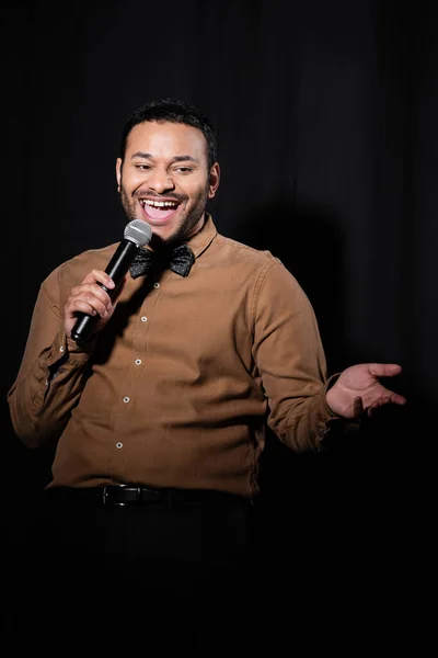 joyful indian comedian in shirt and bow tie holding microphone during monologue on black