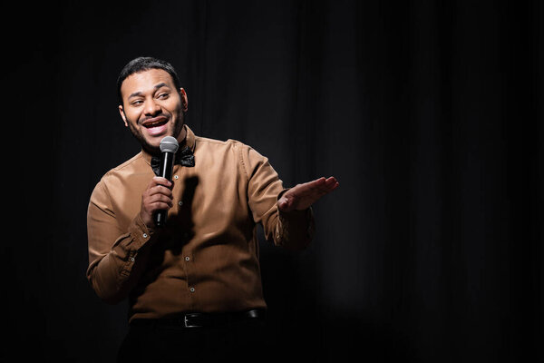 cheerful indian comedian in shirt and bow tie holding microphone during monologue on black 