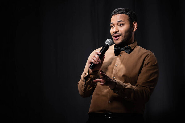 indian comedian in shirt and bow tie gesturing and holding microphone during monologue on black 