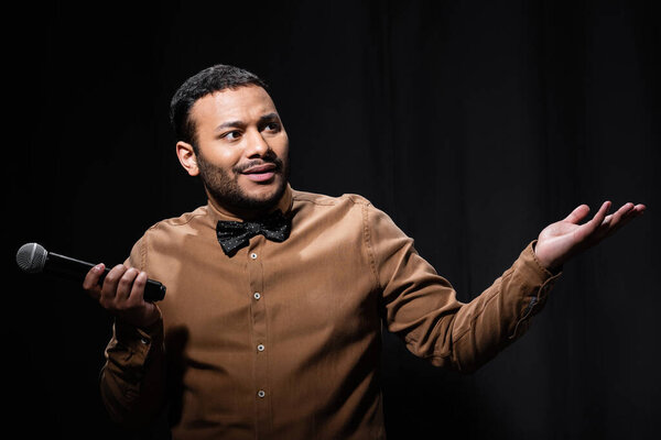 confused indian comedian in shirt and bow tie holding microphone and showing shrug gesture during monologue on black 