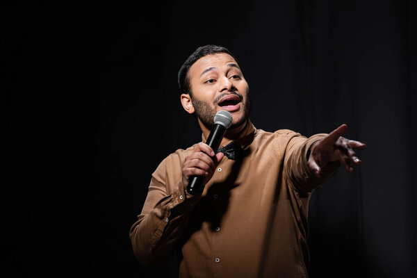 emotional indian comedian with open mouth holding microphone and pointing with finger during monologue on black 