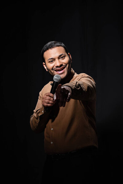 happy indian comedian in shirt and bow tie holding microphone and pointing with finger during monologue on black 