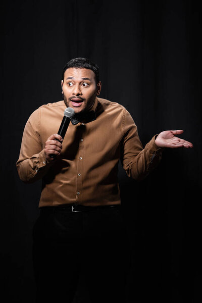 emotional indian comedian in shirt and bow tie holding microphone and gesturing during monologue on black 