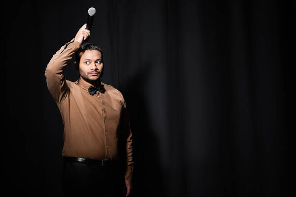 Индийский комик, держащий микрофон на голове на темной сцене на черном 