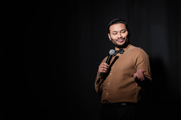 skeptical indian stand up comedian telling joke into microphone on dark stage on black 