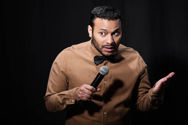 indian stand up comedian gesturing while telling jokes into microphone on dark stage isolated on black 