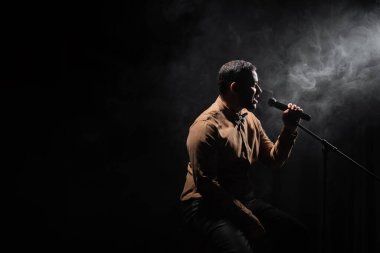 Hintli komedyenin yan görünüşü sandalyede oturup stand-up komedisini siyah üzerine dumanla mikrofona yansıtıyor.