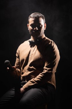 Hintli stand up komedyeni mikrofon tutuyor ve izole bir şekilde oturuyor. 