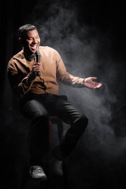 Şaşırmış Hintli komedyen oturuyor ve stand up komedisini siyah üzerine mikrofona yerleştiriyor. 
