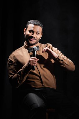 Gülümseyen Hintli komedyen oturur ve siyah üzerine stand-up komedisi yapar. 