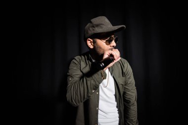 middle east hip hop singer in cap and sunglasses singing into microphone on black 