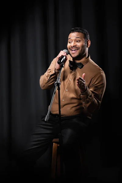 happy indian comedian sitting on chair and performing stand up comedy into microphone on black