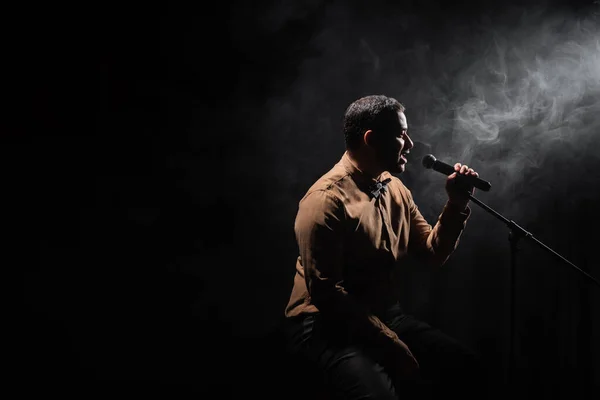 side view of indian comedian sitting on chair and performing stand up comedy into microphone on black with smoke