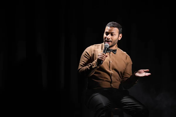 indian comedian sitting and performing stand up comedy into microphone on black