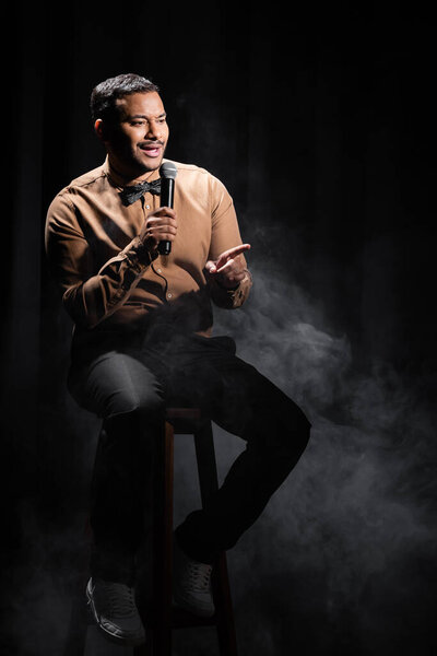 indian comedian sitting and performing stand up comedy into microphone on black with smoke 