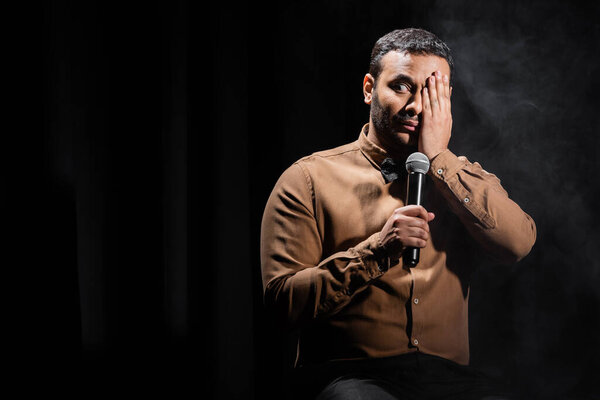 indian comedian covering eye and performing stand up comedy with microphone on black 