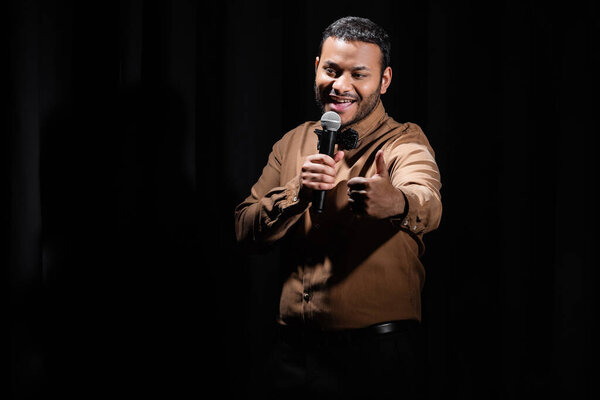 joyful middle east comedian showing like and performing stand up comedy into microphone isolated on black 