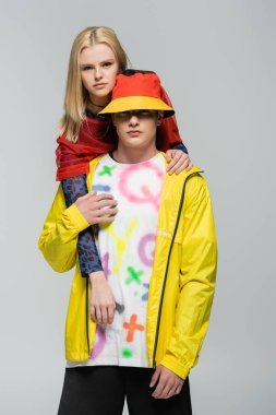 Young blonde woman hugging boyfriend in bright windbreaker and panama hat isolated on grey 
