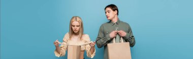 Young man looking at girlfriend with shopping bag isolated on blue, banner 
