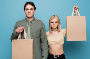 Pretty woman holding shopping bag and touching boyfriend isolated on blue 