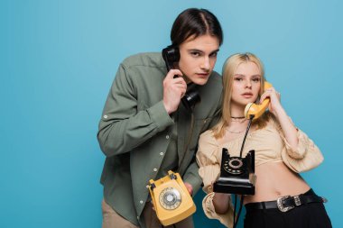 Stylish couple talking on retro telephones and looking at camera on blue background 