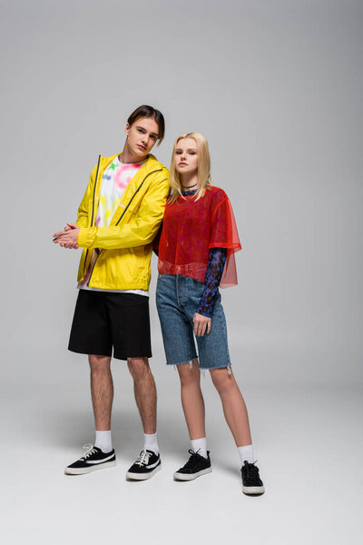Молодой человек в ветровке стоит рядом с блондинкой подружкой на сером фоне