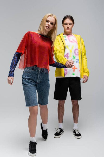 Полная длина молодой женщины в джинсовых шортах держа за руку бойфренда на сером фоне