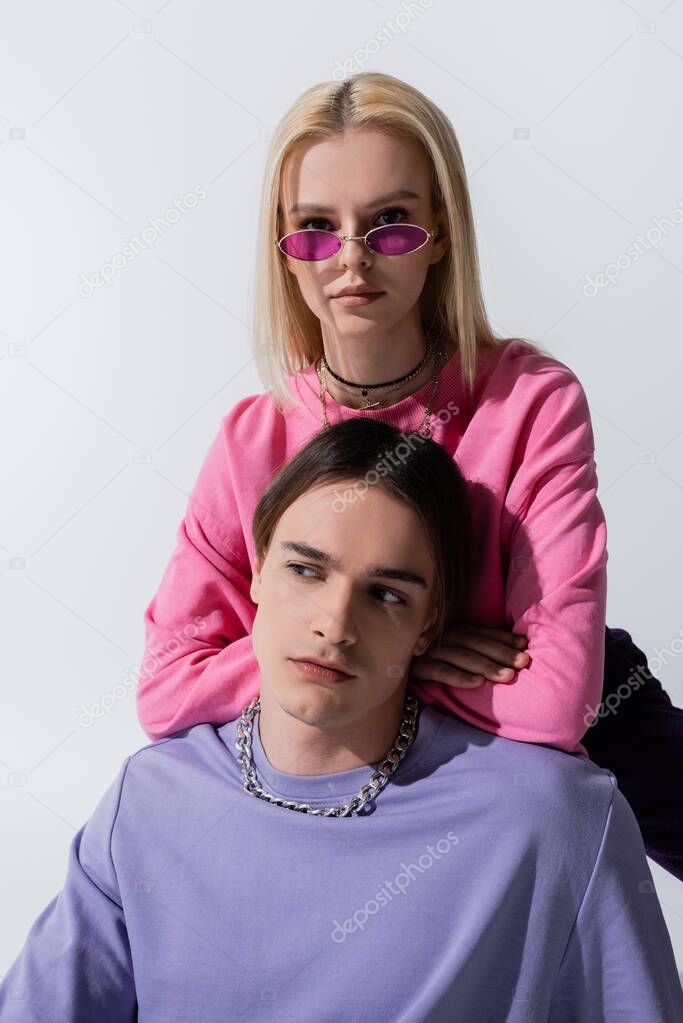 Young blonde model in pink sunglasses looking at camera near boyfriend isolated on grey 