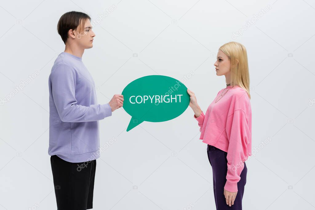 Side view of young couple in sweatshirts holding speech bubble with copyright lettering isolated on grey 