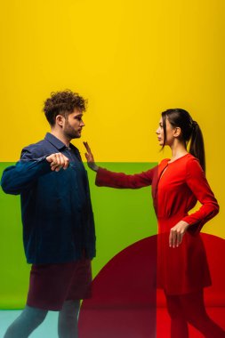 brunette woman showing stop to curly man and holding different shapes glass isolated on yellow