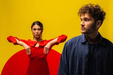 trendy curly man near blurred woman in dress holding round shape glass isolated on yellow