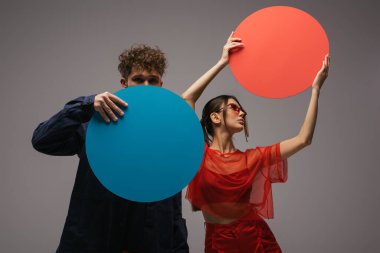 stylish man and woman in trendy blue and red outfits holding round shape carton isolated on grey