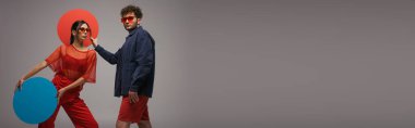 young stylish couple in blue and red outfits holding round shape carton isolated on grey, banner
