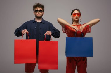 Gri renkli kıyafetler ve güneş gözlüğü takan genç bir çift izole edilmiş alışveriş torbaları taşıyor.