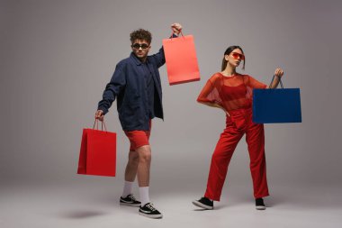 Gri elbiseli, güneş gözlüklü, alışveriş torbaları taşıyan ve gri renkli pozlar veren genç bir çift.