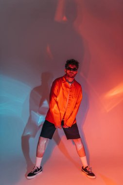 full length of curly and stylish man in sunglasses posing in studio with colorful light