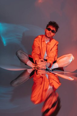 full length of stylish man in sunglasses posing while sitting in studio with colorful light clipart