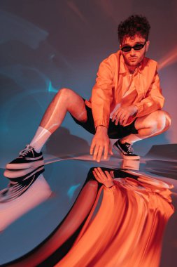 full length of curly and trendy man in sunglasses posing while sitting on grey with colorful light clipart
