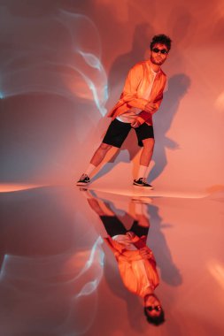 Güneş gözlüklü, tam boy kıvırcık ve modaya uygun bir adam. Bakarken poz veriyor ve kırmızı ışıkta kamera var. 