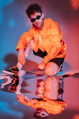 full length of curly young man in sunglasses posing while sitting on blue with red lighting  clipart