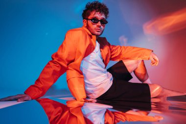 curly young man in sunglasses posing while sitting on blue with red lighting 
