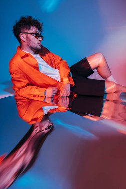 stylish and curly young man in sunglasses posing on blue with red lighting 