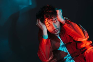 curly and stylish man with closed eyes posing in orange shirt on blue